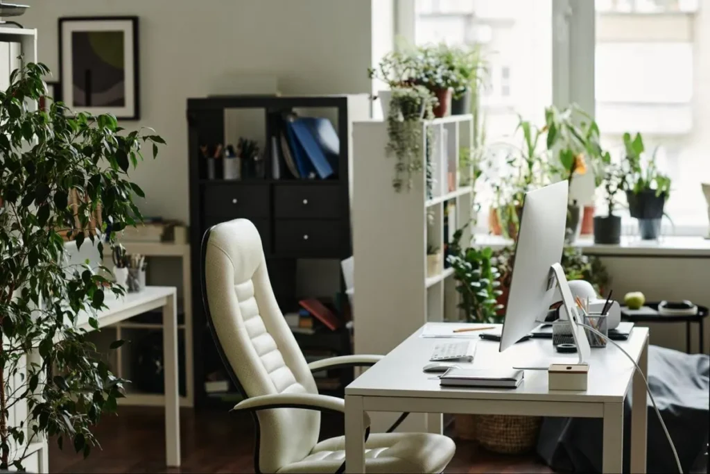Bright home office with a white ergonomic chair, desktop computer, and multiple indoor plants beside a large window for a productive and balanced workspace