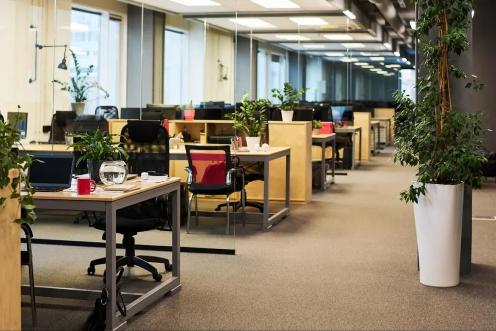 Modern corporate open office with rows of workstations, tall green plants along the aisle, and well-lit, clear walkways supporting employee comfort